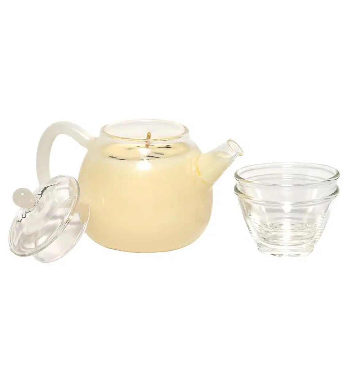 A cozy glass tea pot set filled white soy candle wax and two clear glass tea cups displayed on a rustic wooden round tray, beside a potted green plant on a wooden surface.