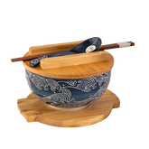 Blue ceramic wave-pattern bowl with bamboo lid, wooden trivet, chopsticks, and matching ceramic spoon.